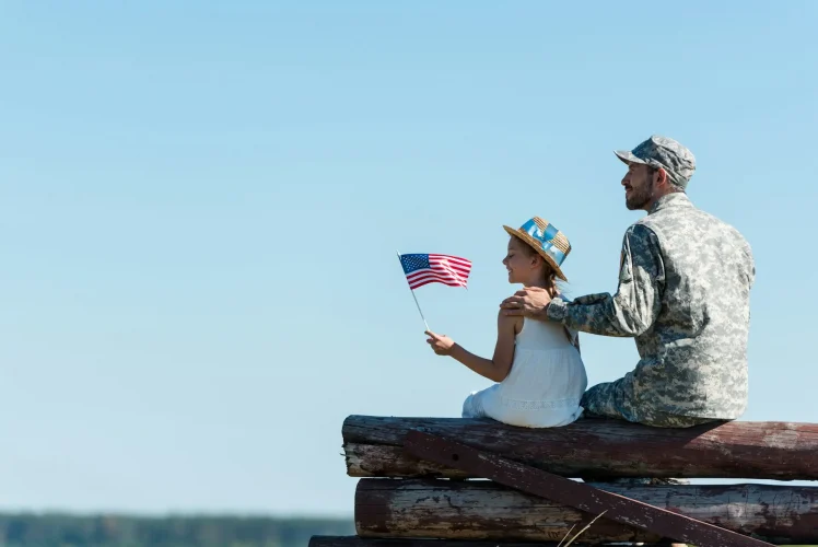 cute child holding american flag near veteran father while sitting in fence